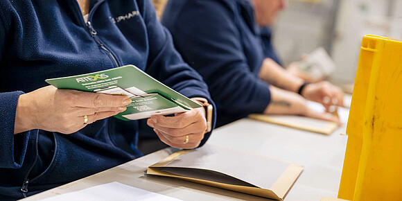 Nahaufnahme der Hände einer Person in blauem Arbeitsfleece, die einen grünen Werbeflyer faltet und in einen Versandumschlag steckt; im Hintergrund arbeiten weitere Personen an Tischen.