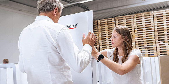 Eine Frau und ein Mann bauen in einer Produktionshalle gemeinsam ein großes, weißes Verkaufsdisplay mit dem Logo „Eucerin“ auf.