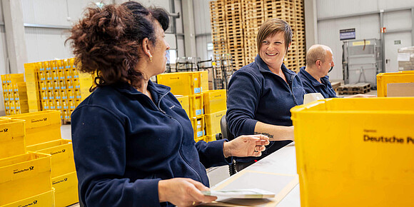 Zwei Mitarbeiter stehen am Tisch im Lager und packen Briefe